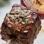 Close-up of a juicy grilled steak cube on a skewer with rosemary seasoning.