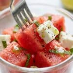 Close-up of a fork lifting a piece of watermelon and feta cheese from a small serving bowl.