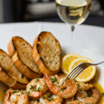 Plated garlic shrimp in a bowl served with toasted baguette slices and a glass of white wine.