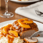 A plated pork chop covered in peach sauce served next to mashed potatoes, with a fork cutting into the meat in a cozy dinner setting.