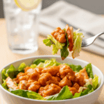 A bowl of buffalo chicken salad on a lunch table, with a fork lifting a bite of chicken and lettuce, set against a casual background.