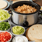 A family-style taco bar setup with a slow cooker of shredded chicken at the center, surrounded by bowls of toppings and tortillas, with a hand building a taco.