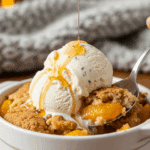 A bowl of warm slow-cooker peach cobbler topped with a melting scoop of vanilla ice cream, served in a cozy setting with a spoon lifting a bite.