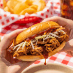 Hands holding a messy pulled pork sandwich at a backyard BBQ, with potato chips and a cold drink in the background.