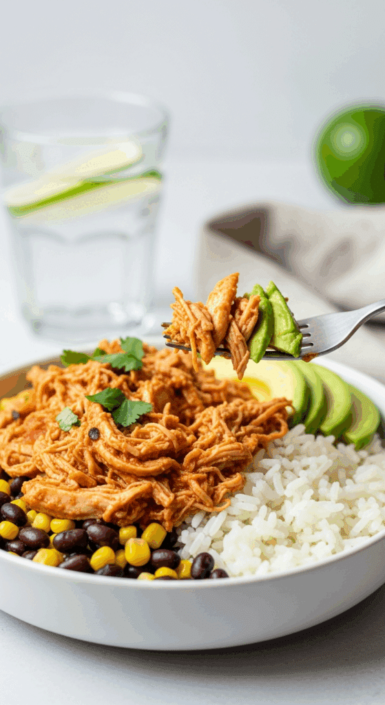 A burrito bowl filled with rice, beans, corn, and salsa chicken, with a fork lifting a bite of chicken and avocado.