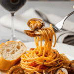 A plate of spaghetti with red clam sauce, with a fork twirling pasta and a clam, served next to a piece of crusty bread for dipping.