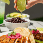 A hand squeezing lime juice over spicy lime chicken tacos filled with avocado and pickled onions, set on an outdoor patio table.