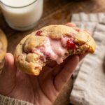 A close-up of a hand holding a bitten Strawberry Cheesecake Cookie, showing the soft, gooey cream cheese and strawberry filling.