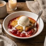 A bowl of warm strawberry cheesecake cake served with a scoop of melting vanilla ice cream.