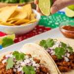 Soft tacos filled with shredded salsa beef being garnished with lime juice, onions, and cheese in a festive dining setting.