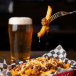 A pub basket filled with chili fries, with a fork lifting a cheesy fry, set against a background of a beer and a sports bar environment.