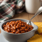 A bowl of thick vegan baked beans served beside a slice of cornbread on a wooden table in a cozy setting.
