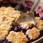 Close-up of a spoon cutting into a fluffy cobbler biscuit and blackberry filling.