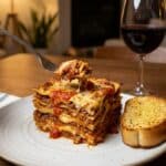 A tall, plated slice of lasagna served with garlic bread and red wine at a cozy dinner table.