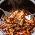 Close-up of forks shredding tender BBQ chicken in a slow cooker.