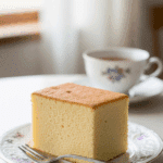 A plain slice of fluffy sponge cake served on a porcelain plate with a fork, next to a cup of tea in a gentle afternoon light setting.