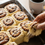 Hand pulling a soft banana cinnamon roll from a baking dish.