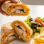 A hand holding a crispy cheesy garlic chicken wrap on a plate with salad and chips.