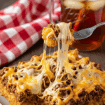 A serving of Cowboy Casserole on a plate with a fork, showing the cheesy texture and tater tots, served with a drink in the background.