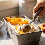 An ice cream scoop taking a serving from a loaf pan filled with homemade no-churn peach ice cream.