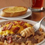 A bowl of creamy bacon cheeseburger pasta plated for dinner with a fork, set on a wooden table with garlic bread in the background.
