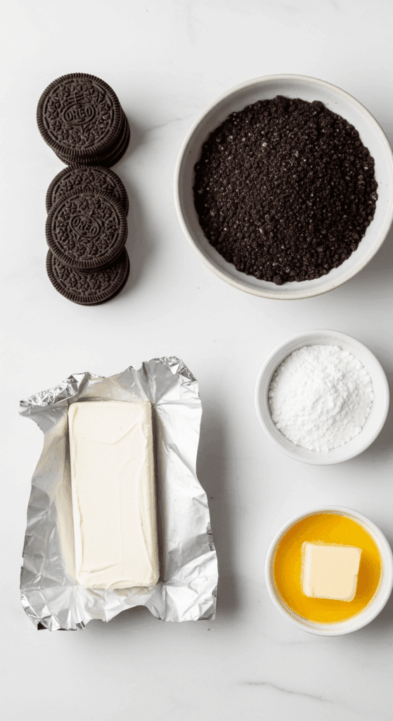Overhead view of ingredients for Oreo Dessert Sushi on a marble counter: whole Oreos, crushed Oreos, cream cheese block, powdered sugar, and melted butter.