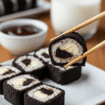 A slice of Oreo cheesecake sushi being lifted by chopsticks from a plate, with a bowl of chocolate sauce in the background.