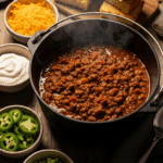 Large Dutch oven of Cowboy Chili with bowls of toppings and cornbread on a table.