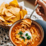 Person holding a warm bowl of creamy chicken enchilada soup with a spoon and tortilla chips.